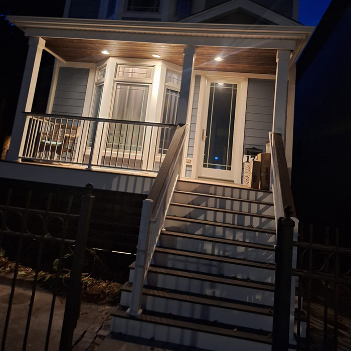 Porche bien iluminado de una casa por la noche. Unos escalones conducen a una puerta de cristal, flanqueada por una barandilla blanca. La cálida iluminación crea un ambiente acogedor y sugerente.