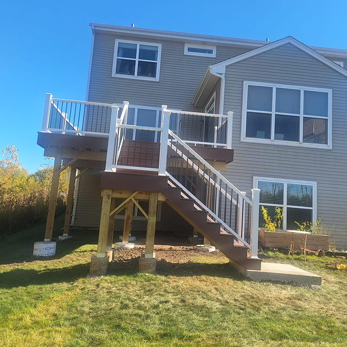 Una casa de dos pisos con revestimiento color canela cuenta con una terraza elevada de madera con barandillas y escaleras blancas. Se alza contra un cielo azul claro y un jardín con césped.