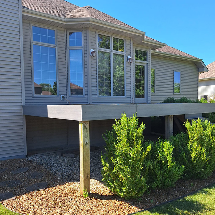 Una casa beige con grandes ventanales de rejilla y techo marrón. Tiene una terraza elevada de madera sin barandillas, rodeada de arbustos verdes.