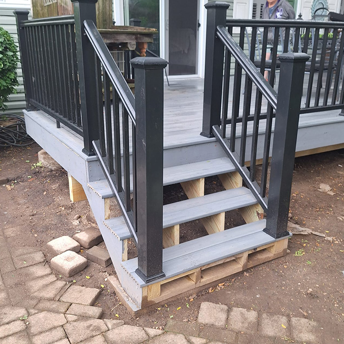 Terraza de madera gris con barandillas negras y escaleras sostenidas por palés a la vista. Un patio de ladrillo y tierra de jardín rodean la zona, creando un ambiente inacabado.