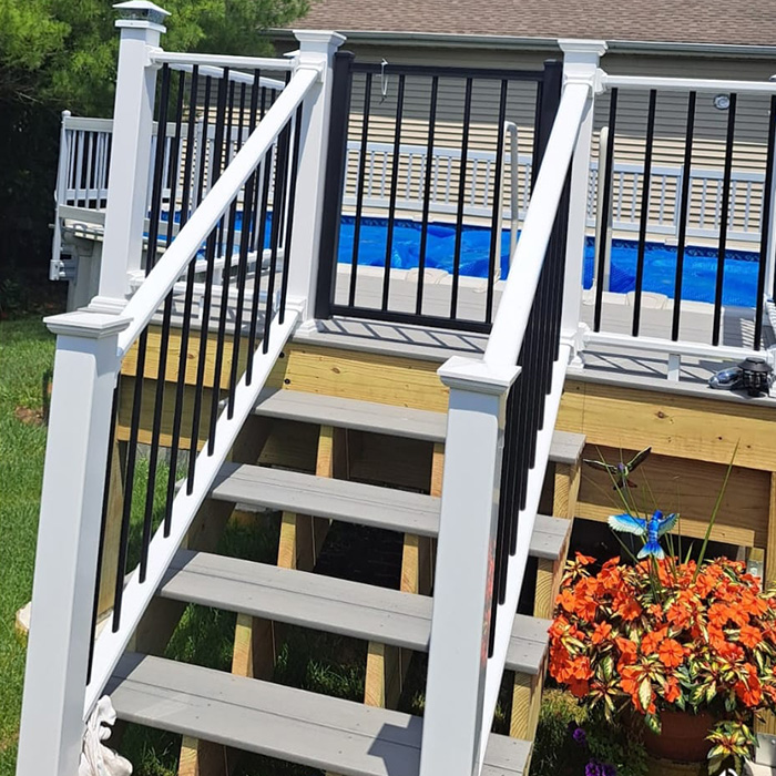 Unas escaleras exteriores de madera con barandillas blancas y negras conducen a una terraza con una piscina cubierta azul. Cerca, unas flores de color naranja brillante le dan un toque vibrante.